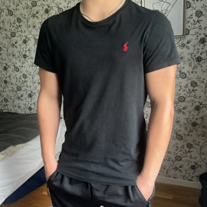Svart t-shirt från Ralph Lauren - Svart t-shirt från Ralph Lauren med klassisk röd broderad logga på bröstet. T-shirten har rund halsringning och korta ärmar. Tillverkad i mjuk bomull och har en normal passform som sitter snyggt på kroppen. Storlek M men sitter bra på mig som använder S vanligtvis! Bara att skriva vid minsta fundering