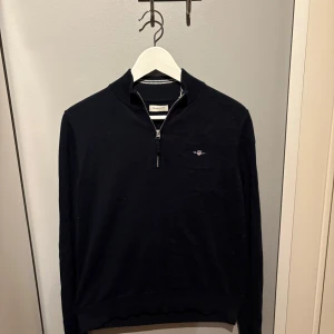 Svart half zip tröja från GANT - Snygg Svart stickad tröja från GANT med half zip och broderad logga på bröstet. Tröjan har ribbade muddar och krage med vita ränder på insidan. Perfekt för dig som gillar klassisk och stilren look. Nypris: 1099kr
