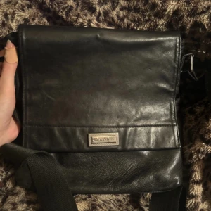 Svart axelväska i äkta lammnappa - Snygg svart axelväska från New Style i äkta lammnappa. Väskan har ett stilrent och klassiskt utseende med lock, justerbar axelrem och flera fack med dragkedja. Perfekt för dig som vill ha en praktisk och tidlös väska med plats för det viktigaste.