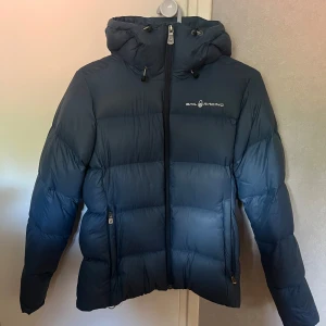 Mörkblå dunjacka från Sail Racing - Säljer en mörkblå dunjacka från Sail Racing med huva och dragkedja framtill. Jackan har två fickor med dragkedja och logga på bröstet samt bak på huvan. Perfekt för kalla dagar och riktigt snygg passform.