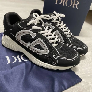 Dior skor - Säljer ett par svarta Dior sneakers med stora vita CD-loggor på sidorna och vita snören. Skorna har en chunky vit och svart sula och är tillverkade i mesh och syntetmaterial. Snygg kontrast mellan svart och vitt, perfekt för dig som gillar statement sneakers.