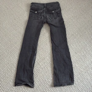Svarta bootcut jeans med fickdetaljer - Snygga svarta jeans med bootcut passform och coola fickor med lock och silverknappar bak. Jeansen har klassisk femficksdesign, bälteshällor och är tillverkade i mjukt denimtyg. Perfekta för dig som gillar en avslappnad men trendig look.