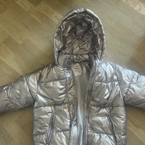 Silver pufferjacka från Lefties - Säljer en snygg silverfärgad pufferjacka barn från Lefties. Jackan har huva, dragkedja och två fickor med dragkedja framtill.