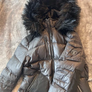 Svart pufferjacka Rockandblue med fuskpäls - Snygg svart pufferjacka från Rockandblue med stor huva och fuskpäls. Jackan har flera dragkedjor, praktiska fickor och en patch på ärmen. Perfekt för kalla dagar med sin vadderade design och coola detaljer. Knappt använd därav priset. Liten i storleken. (priset går att diskutera)