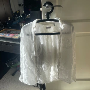 Vit linneskjorta från GANT - Fräsch vit skjorta från GANT i luftig linnemix, perfekt för varma dagar. Klassisk krage, knappstängning framtill och bröstficka med diskret broderad logga. Skjortan har lång ärm och en lätt, transparent känsla som ger en chill vibe.