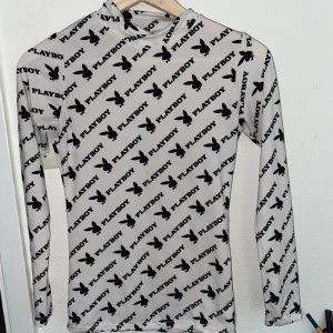 Vit långärmad topp från Playboy - Snygg vit långärmad topp från Playboy med svart logotyp- och kaninmönster över hela plagget. Topp med hög hals och tight passform, perfekt för att sticka ut. Materialet känns stretchigt och lätt.