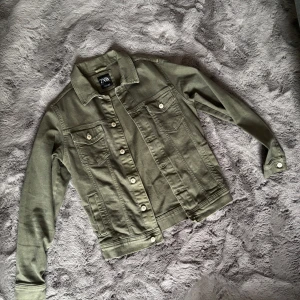 Olivgrön jeansjacka från Zara - Snygg olivgrön jeansjacka från Zara med klassisk krage, vita knappar och två bröstfickor med lock. Jackan har lång ärm och raka linjer, perfekt för en cool och avslappnad stil.