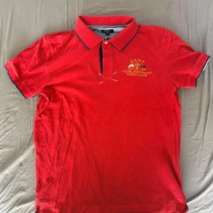 Röd GANT  pikétröja S - Snygg röd pikétröja från GANT med broderad Yacht Club-logga i gult på bröstet. Klassisk krage med mörkblå kant och knappslå. Tillverkad i bomull, kortärmad och med coola detaljer som broderade flaggor. Perfekt för dig som gillar preppy stil.