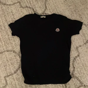Svart t-shirt från Moncler - Svart t-shirt från Moncler med klassisk passform och rund hals. T-shirten har korta ärmar och en liten broderad Moncler-logga på bröstet. Tillverkad i mjuk bomull som känns skön mot huden. Perfekt för en clean och stilren look.