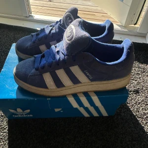 Adidas Campus blå sneakers 43 1/3 - Säljer ett par Adidas Campus sneakers i blå mocka med vita klassiska stripes och logga på plösen. Skorna har platt sula, rund tå och snörning. Perfekta för dig som gillar retrovibbar och streetstyle. Storlek 43 1/3 (EU).