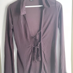 Brun långärmad topp med snörning - Säljer en brun långärmad topp med djup v-ringning och snygg snörning framtill. Tunn och lätt i materialet, perfekt för att styla lager på lager. Krage upptill ger en extra touch. Passar dig som gillar trendiga detaljer och en lite edgy look.