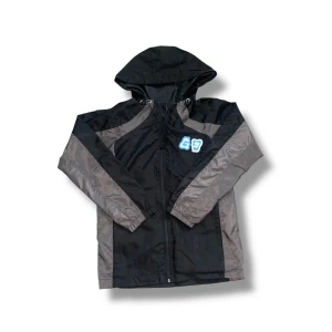 Svart windbreaker från GravityGrab - Snygg svart och grå vindjacka från GravityGrab med huva och dragkedja. Jackan har coolt blåvitt tryck på bröstet och stort graffiti-inspirerat tryck på ryggen med texten 'The Only Destination'. Perfekt för dig som gillar streetwear vibes. Först i värden att få denna windbreakern, säljs ej längre.