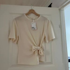 Vit topp med knyt från H&M - Säljer en snygg vit/beige topp från H&M i mjuk bomull med kort ärm och rund hals. Toppen har en knytdetalj framtill som man kan knyta hur man vill. Tröjan är i nytt skick med prislappen kvar och därmed aldrig använd💕💕