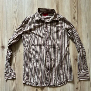 Randig skjorta från Hugo Boss S - Säljer en snygg skjorta från Hugo Boss i storlek S. Skjortan har beige botten med smala ränder i brunt, rosa och vitt. Klassisk krage, långa ärmar och knäppning framtill. Perfekt för dig som gillar stilrena och trendiga plagg.