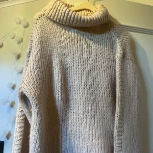 Beige stickad polotröja Gina Tricot - Mysig beige stickad polotröja från Gina Tricot med grovstickad struktur och bred krage. Tröjan har lång ärm och dekorativa detaljer i guld längs sidorna. Perfekt för kyliga dagar och enkel att styla till jeans eller kjol.