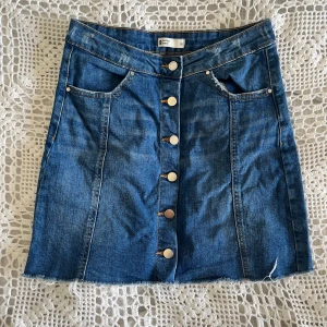 Blå jeanskjol med knappar från Gina Tricot -  Säljer en blå jeanskjol från Gina Tricot med snygga silverfärgade knappar framtill. Kjolen har två fickor fram och markerade sömmar.  stretchigt material vilket för den perfekt att använda som topp om man vill vara kreativ, se sista bilden.