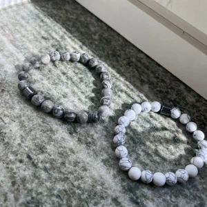 Två marmorerade pärlarmband - Säljer två stilrena armband med runda pärlor i marmorerad design. Ett armband är vitt med gråa ådror och det andra är grått med mörkare marmorering. Båda har en metallisk cylinderpärla som detalj. Perfekta accessoarer för en clean och modern look. 2 för 60kr