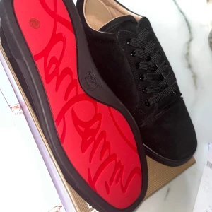 Christian Louboutin skor  - Snygga svarta sneakers från Christian Louboutin i mocka med ikonisk röd sula och diskret logga på sidan. Skorna har svarta snören och en stilren, minimalistisk design som sticker ut med den klassiska röda undersidan. Perfekta för dig som vill ha något exklusivt och trendigt.