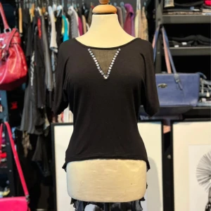 Svart topp med mesh och pärldetaljer - Svart topp från Little Mistress med korta ärmar och transparent meshparti framtill, dekorerad med pärlor i V-form. Baktill har toppen en längre, transparent meshdel som ger en cool och unik look. Storlek S/M