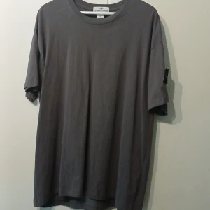 Mörkgrå t-shirt från Stone Island 4XL - Säljer en mörkgrå t-shirt från Stone Island i storlek 4XL. T-shirten är i bomull och har klassisk rund hals samt korta ärmar. På vänster ärm sitter den ikoniska Stone Island-patchen med broderad logga i gult och grönt. Perfekt för dig som gillar streetwear och oversized stil.