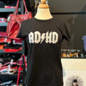 Svart t-shirt med ADHD-tryck - Svart t-shirt i 100% bomull med  ADHD-tryck framtill i vit/grå stil som påminner om rockbandet AC/DC. Modellen är klassisk med rund hals och korta ärmar.
