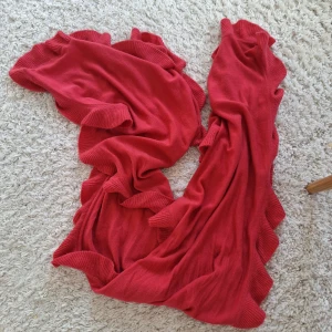 Röd stickad halsduk med volangkant - Säljer en snygg röd stickad halsduk med bred volangkant längs hela sidorna. Korallrosa. Halsduken är mjuk och har en härlig färg som verkligen poppar. Perfekt accessoar för att lyfta din outfit under kyliga dagar. 200 cm rakt över