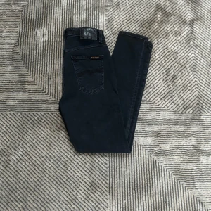 Svarta jeans från Nudie Jeans - Svarta skinny jeans från Nudie Jeans med klassisk femficksdesign och hög midja. Jeansen är tillverkade i stretchig bomullsdenim för en tight och bekväm passform. Snygga detaljer med svarta sömmar och Nudie-logga på bakfickan.