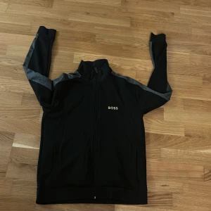 Svart ziptröja från BOSS med grå detaljer - Svart ziptröja från BOSS med grå ränder längs ärmarna och diskret BOSS-logga på bröstet. Tröjan har hög krage, hel dragkedja och två fickor framtill. Perfekt för en chill och stilren look. Ny pris 1200