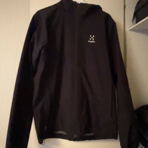 Svart vindjacka från Haglöfs - Svart vindjacka från Haglöfs med minimalistisk design och diskret logga på bröstet. Jackan har huva och långa ärmar, perfekt för blåsiga dagar. Tillverkad i lätt polyester som känns smidig och enkel att bära.