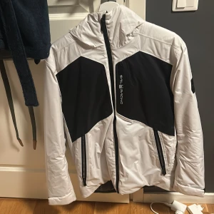 Vit och svart Sail Racing jacka - Snygg vit och svart jacka från Sail Racing med huva och dragkedjor framtill. Jackan har sportig design med svarta paneler på bröstet och ryggen, samt flera praktiska fickor med dragkedja. Perfekt för dig som gillar stilren och skön vinterjacka . Använd 2-3 gånger. 