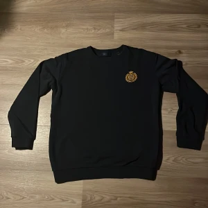 Svart sweatshirt från GANT med emblem - Svart sweatshirt från GANT med rund hals och ribbade muddar. På bröstet finns ett snyggt broderat guldemblem som ger en exklusiv känsla. Perfekt för dig som gillar stilrena och klassiska plagg med en twist.