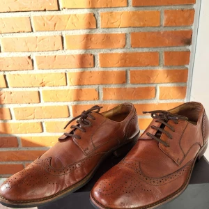 Bruna skinnloafers från Dahlin eUR 43 - Snygga bruna loafers från Dahlin i äkta skinn med klassisk broguedetalj och snörning. Skorna har rund tå, låg klack och dekorativa perforeringar längs kanterna. Perfekta för dig som gillar stilrena och tidlösa skor.
