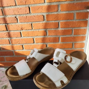 Vita sandaler från Rieker, storlek 37 - Säljer ett par vita sandaler från Rieker i storlek 37. De har breda remmar i skinn, snyggt spänne i brunt och en bekväm platt sula. Perfekt för dig som gillar enkel och stilren design med fokus på komfort. Sulan är brun och har dekorativa sömmar runt kanten.