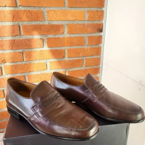 Bruna loafers i skinn med dekorativ söm EUR 42 - Snygga bruna loafers från AR i äkta skinn med dekorativa svarta sömmar över vristen. Skorna har låg klack och rund tå, klassisk design och platt sula. Perfekta för dig som gillar stilrena och tidlösa skor med lite extra detalj.
