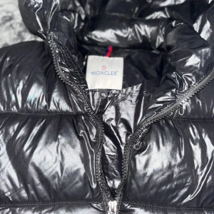 Svart Moncler pufferjacka med huva - Säljer en svart glansig pufferjacka från Moncler med huva och dragkedja. Jackan har klassisk Moncler-logga på ärmen, två fickor med dragkedja och quiltad design. Perfekt för kalla dagar och riktigt snygg streetstil. storlek S