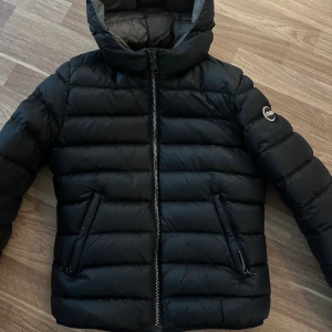 Svart pufferjacka från Colmar - Snygg svart pufferjacka från Colmar med huva och dragkedja framtill. Jackan har quiltad design, två fickor med dragkedja och Colmar-logga på ärmen. Perfekt för kalla dagar och ger en clean, sportig look.