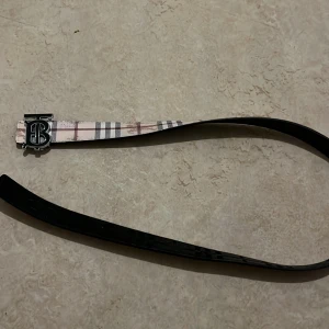 Burberry rutigt bälte med silverdetalj - Snyggt bälte från Burberry med klassiskt rutigt mönster i beige, svart och rött på ena sidan och helsvart på andra. Spännet är i silver med Burberrys ikoniska TB-logga. Perfekt accessoar för att lyfta din outfit.