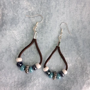 Handgjorda örhängen  - Unika örhängen med mörkbrunt läderimiterat mockaband och iriserande pärlor i vitt, blått, turkost och samt en grå i mitten . Pärlorna är trädda på bandet i en droppformad design och ger en cool boho-vibe. Perfekta för dig som vill sticka ut med färg och form. Öronkroken är nickelsäker och aldrig använda.