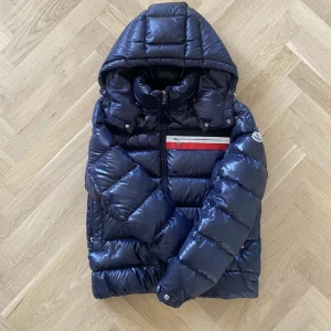 Mörkblå Moncler dunjacka med huva - Snygg mörkblå dunjacka från Moncler med glansig finish, avtagbar huva och dragkedja framtill. Jackan har en vit och röd randdetalj på bröstet, Moncler-logga på ärmen och quiltad design. Perfekt för kalla dagar och riktigt stilren look.