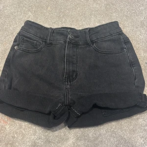 Svarta jeansshorts från UNIQ YOUNG - Svarta jeansshorts från UNIQ YOUNG med klassisk femficksdesign och uppvikta benslut. Shortsen har bälteshällor och knappstängning framtill. Perfekta för en chill sommarlook och lätta att matcha med allt.