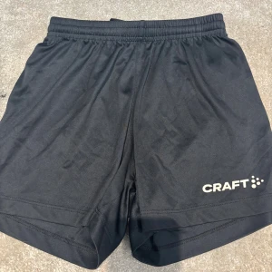 Svarta fotbollsshorts från Craft - Svarta fotbollsshorts från Craft i juniorstorlek 134-140. De är tillverkade i ett lätt och svalt syntetmaterial, har elastisk midja med snörning och en enkel, klassisk design med logga på benet. Perfekta för träning eller idrottslektioner.