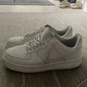 Nike Air Force 1 vita sneakers herr - Klassiska Nike Air Force 1 sneakers i helvitt skinn med lågt skaft och rund tå. Skorna har perforerad tåbox, snörning och den ikoniska swoosh-loggan på sidorna. Perfekta för dig som gillar en clean och tidlös stil.