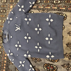 Blå Louis Vuitton stickad tröja - Säljer en blå stickad tröja från Louis Vuitton med ikoniskt LV-mönster i vitt över hela tröjan. Tröjan har rund halsringning och långa ärmar. Perfekt statement piece för dig som vill sticka ut med lyxig streetstyle.