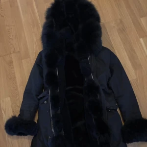 Svart parkajacka med pälsdetaljer - Säljer en svart parkajacka med fluffiga pälsdetaljer på huva, ärmslut och insida. Jackan har dragkedja och fickor, den är riktigt varm. Det är en Fatima jacka och köptes för 3999