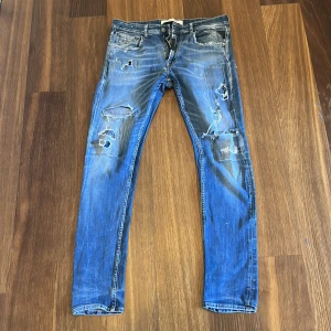 Blå Replay jeans med slitningar - Säljer ett par blå jeans från Replay med coola slitningar och trasiga detaljer på benen. Jeansen har klassisk femficksdesign, smal passform och är tillverkade i jeansmaterial. Perfekta för dig som gillar en avslappnad och trendig look.