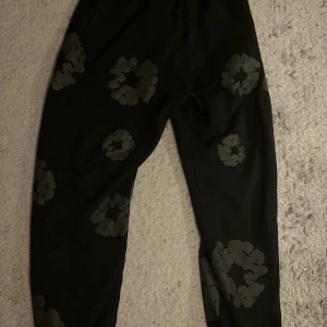 Svarta mjukisbyxor Denim Tears S - Svarta mjukisbyxor från Denim Tears med unika blommönster i mörkgrått över hela byxorna. Byxorna har elastisk midja och är tillverkade i mjukt bomullsmaterial. Perfekt för chill dagar och streetwear vibes.