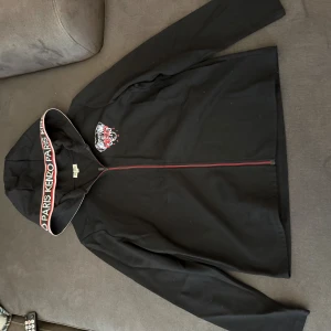 Svart Kenzo hoodiejacka med tryck - Svart hoodiejacka från Kenzo med ikoniskt tigertryck och logga på bröstet. Huva med bredt band där det står 'KENZO PARIS' i vitt och rött. Jackan har hel dragkedja framtill och är tillverkad i mjuk bomull. Perfekt för en streetstyle-look.