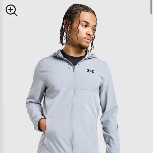 Ljusgrå vindjacka från Under Armour - Snygg ljusgrå vindjacka från Under Armour med huva och dragkedja hela vägen. Jackan har två sidofickor med dragkedja och en stilren logga på bröstet. Perfekt för dig som gillar sportig och clean stil.