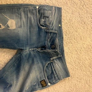 G-Star Raw blå slitna jeans low waist  - Säljer ett par blå jeans från G-Star Raw med slitningar och snygga detaljer. Jeansen har dragkedja på bakfickan, klassisk femficksdesign och en cool tvättad look. Perfekta för dig som gillar streetstyle och vill ha något unikt i garderoben. Low waist 