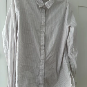 Vit randig skjorta med knappar - Klassisk vit skjorta med tunna grå ränder och knäppning framtill. Skjortan har lång ärm och krage, perfekt för en clean och stilren look. Tillverkad i mjuk bomull som känns skön mot huden.